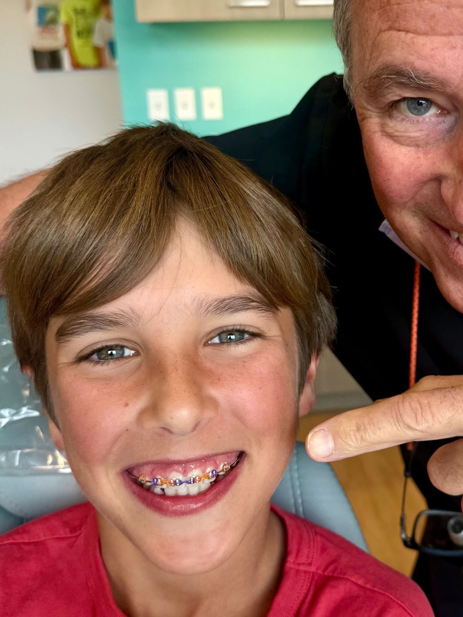 A smiling boy showing his braces with orange and purple bands while Dr. Robert Miller points to his smile