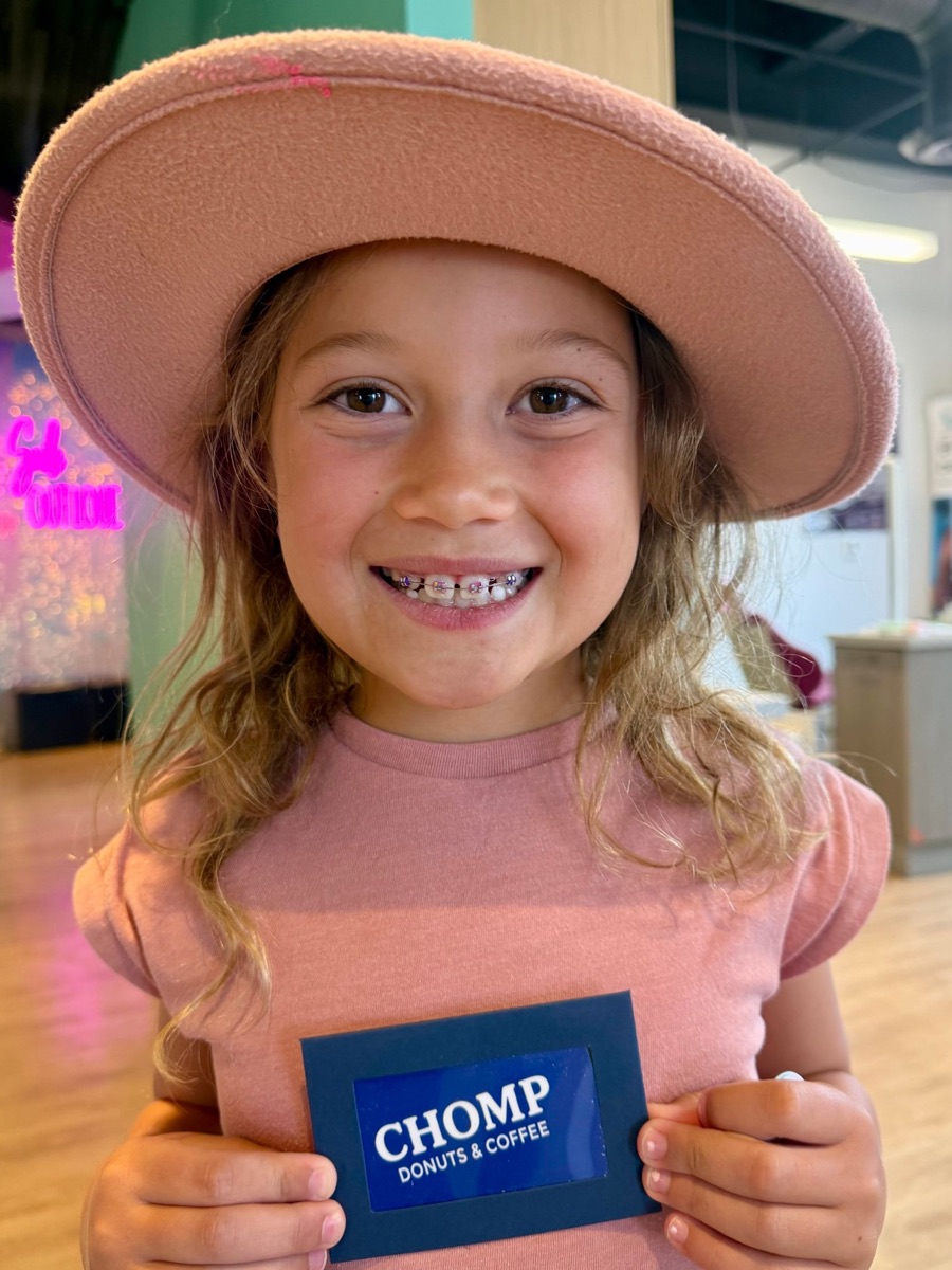 A smiling young girl in a pink hat showing her braces and holding a CHOMP Donuts & Coffee gift card