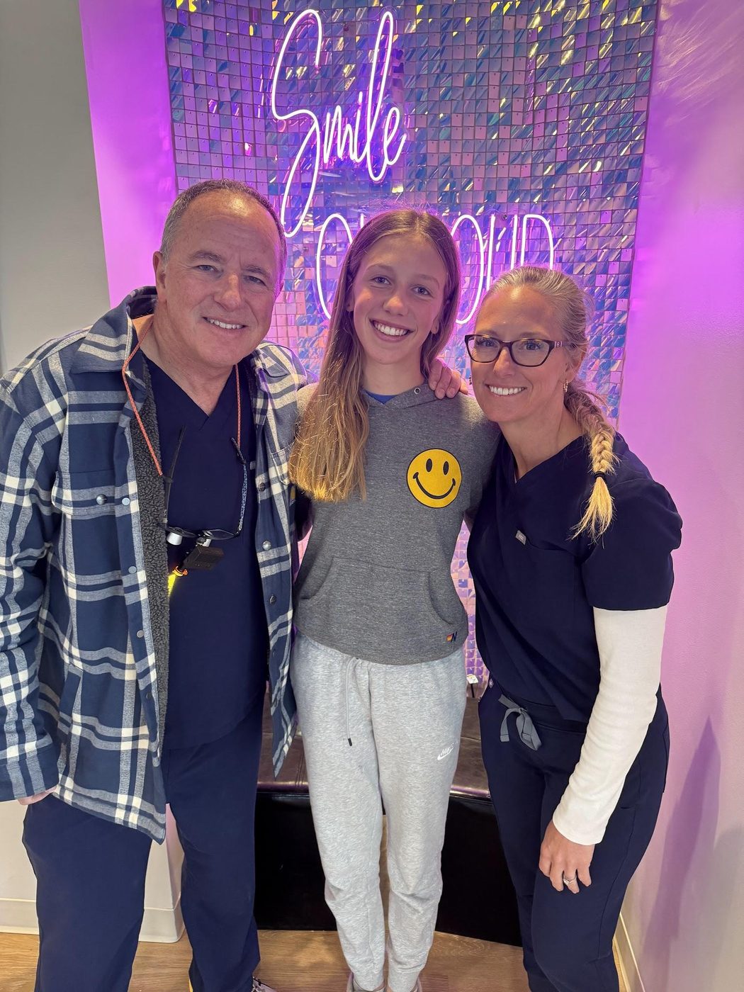 Miller Orthodontics doctors with a smiling patient in front of the office’s neon Smile sign
