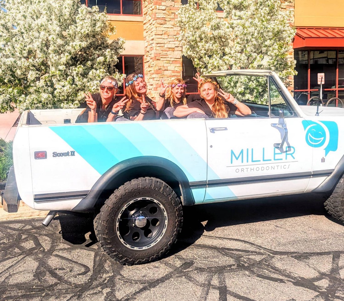 Four Miller Orthodontics team members in flower crowns throwing peace signs from the back of the practice's white Scout II, which carries the Miller Orthodontics logo